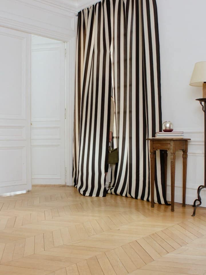 Interior view with striped curtain, side table, books, lamp, and parquet flooring against white walls and doorway in a bright setting.