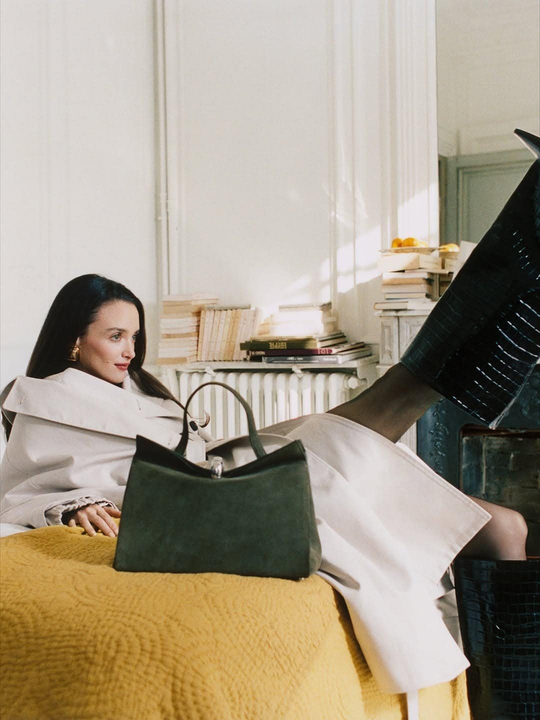 Model seated on a yellow bed with a green handbag in foreground and dark boots raised
