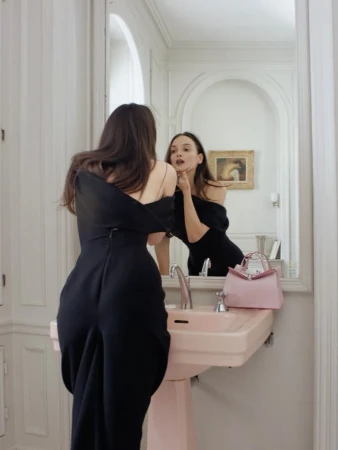 Woman in black off-the-shoulder dress standing before a bathroom mirror with a pink sink and handbag