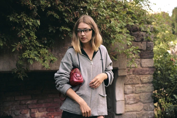 Gigi Hadid in a grey zip hoodie wearing sunglasses holding a red handbag outdoors against stone and foliage.