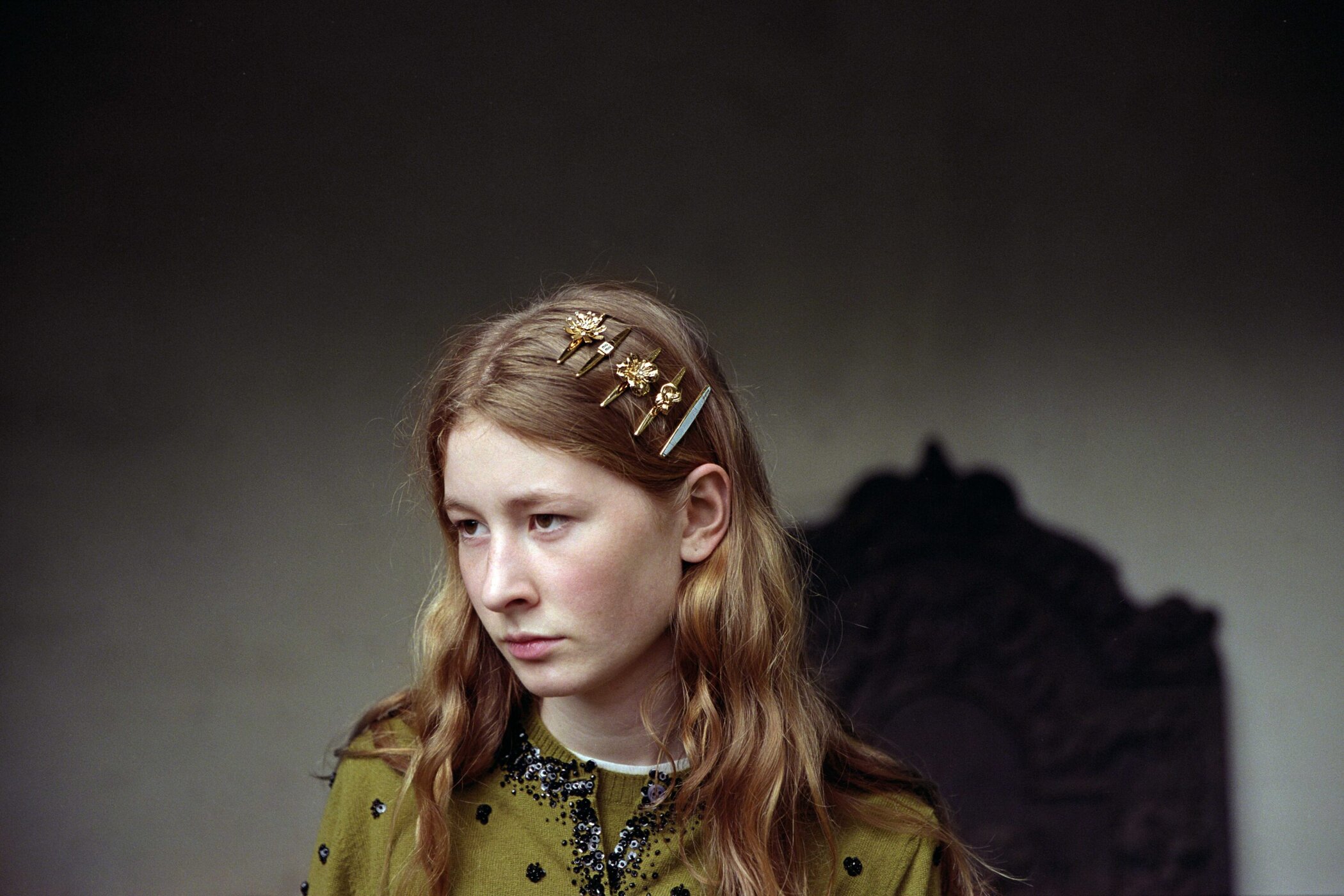 Young woman with blonde hair wearing flower hair clips and a green cardigan, portrait shot indoors with a blurred background.