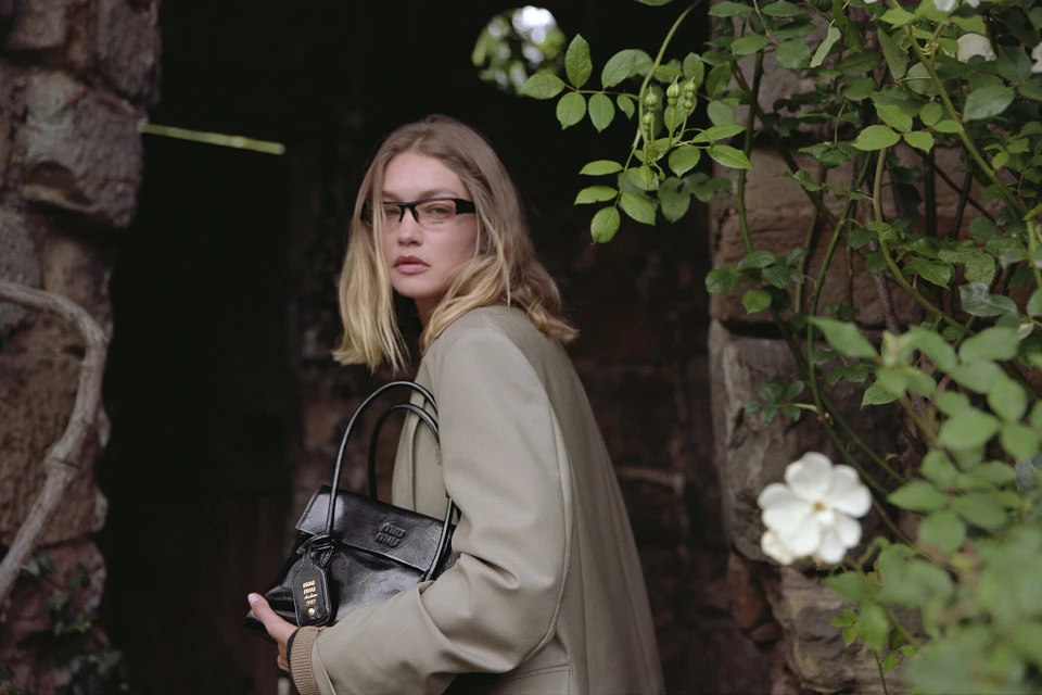 A blonde woman with glasses and a neutral expression is walking out of a stone doorway with a black purse, captured in natural light.