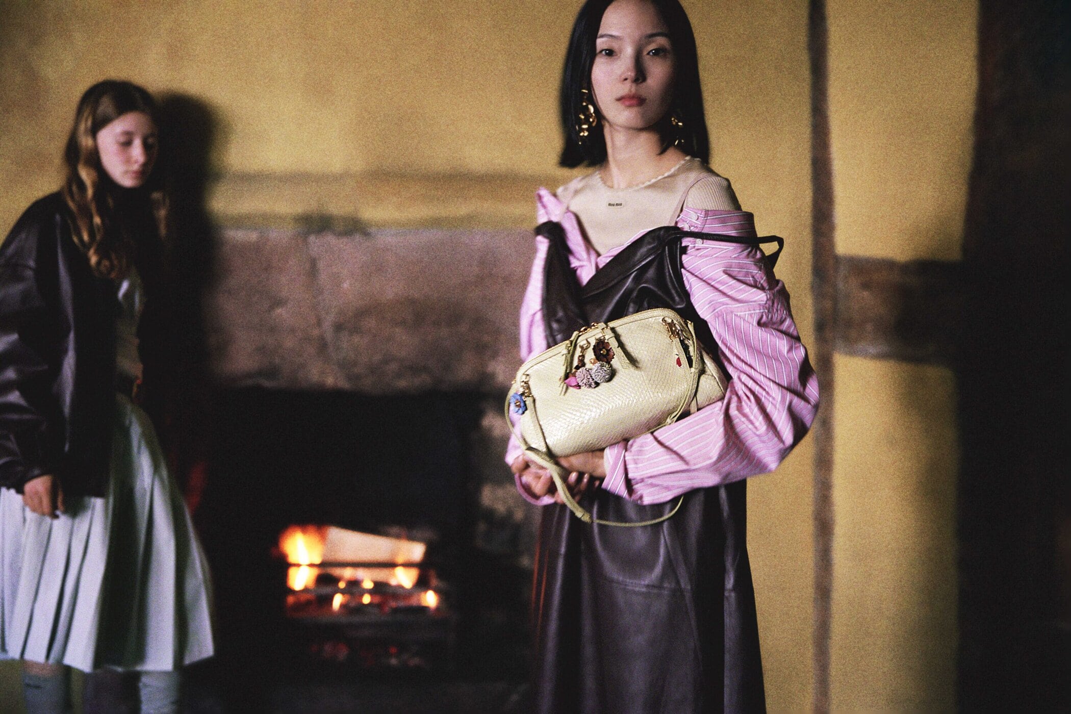 Two young women pose indoors in a dimly lit room with yellow walls. One woman holds a light beige purse. A fireplace is visible in the background.