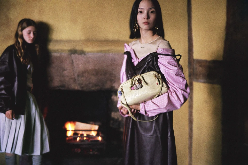 Two young women pose indoors in a dimly lit room with yellow walls. One woman holds a light beige purse. A fireplace is visible in the background.