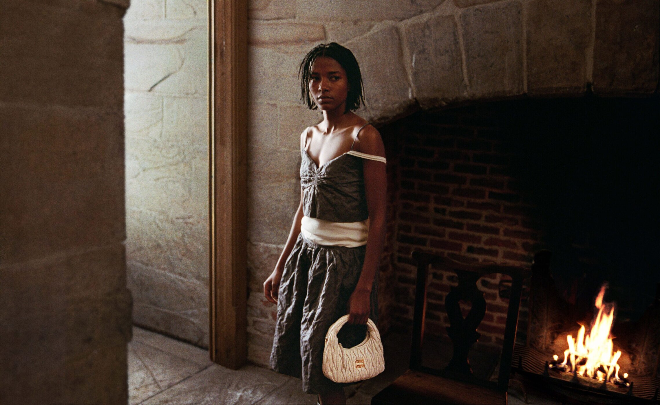 A woman in a dress stands in a stone hallway next to an open fireplace, holding a light-colored purse in her left hand.
