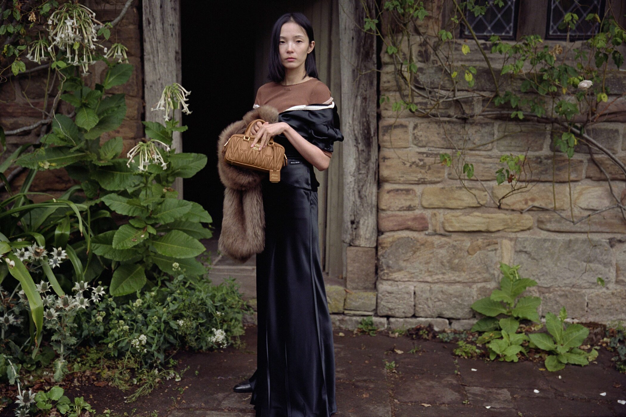 Model wearing dark silk dress and holding tan handbag standing outside historic stone building with lush foliage