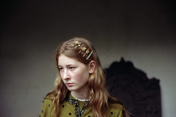 Close-up portrait of a woman wearing an olive green embellished cardigan and gold hair clips against a muted interior background.