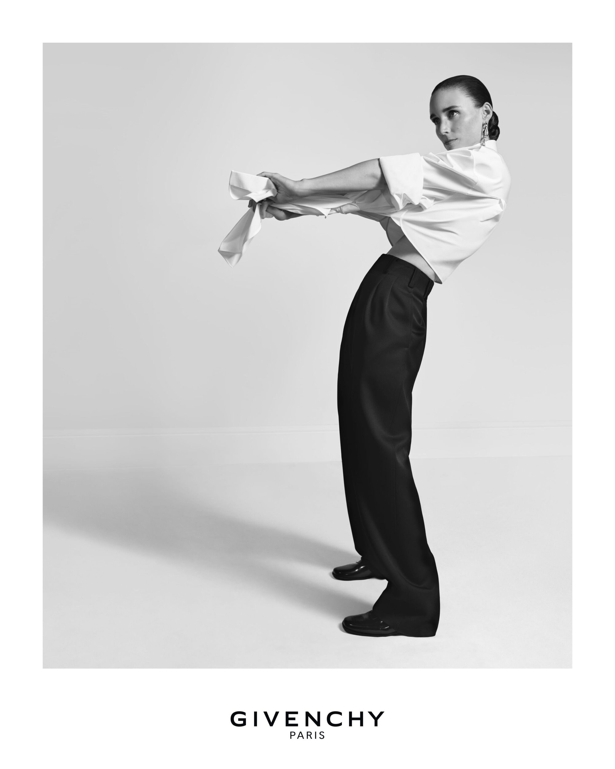Fashion editorial shot in black and white featuring a posed adult female, dressed in a white blouse and black pants, holding white cloth.
