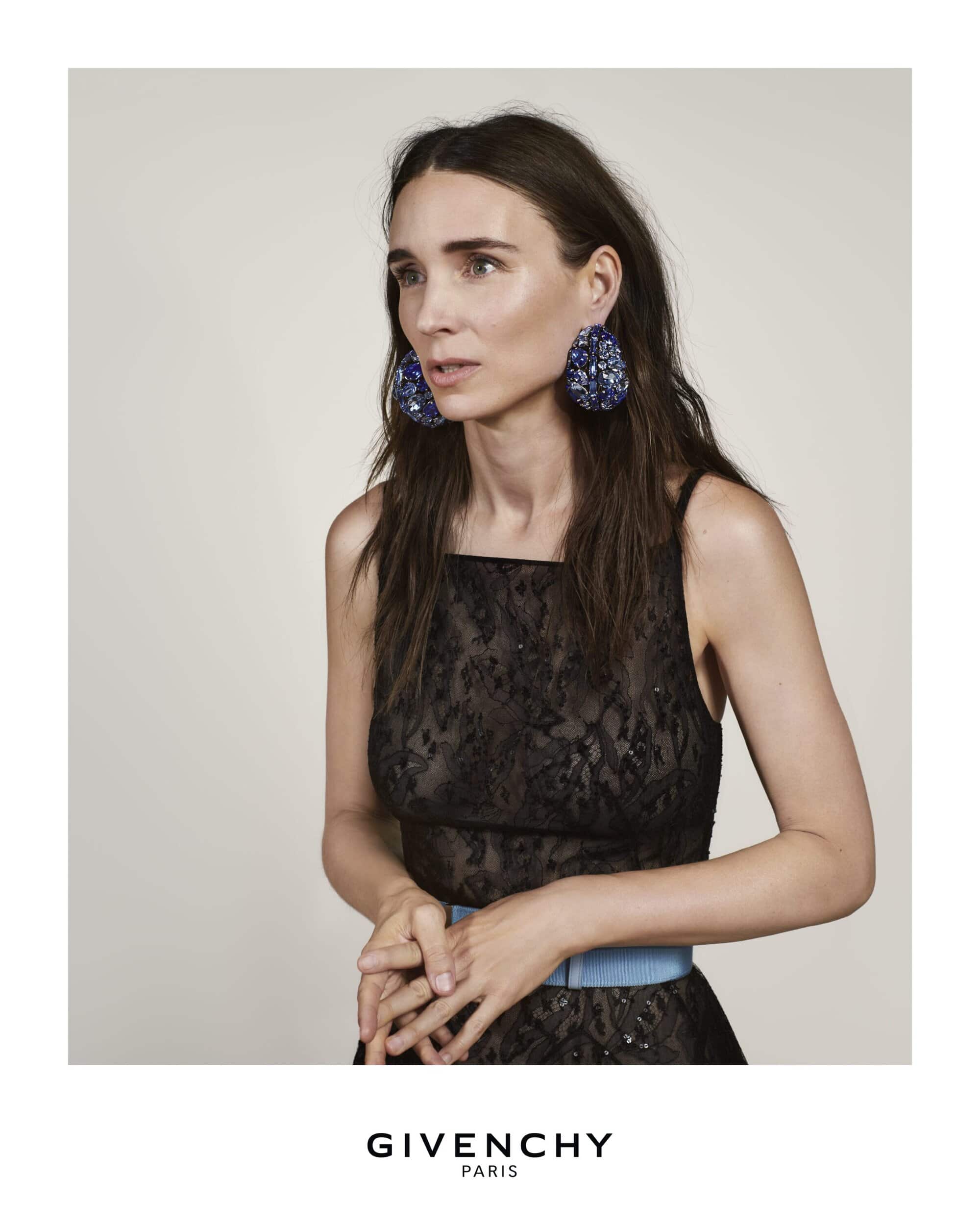 Elegant studio shot of a stylish woman with dark hair and large earrings, wearing a black dress and light blue belt against neutral backdrop.