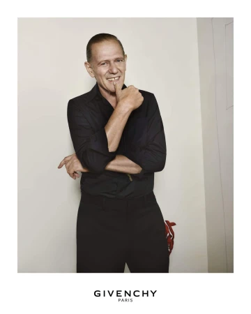 Man in black shirt and pants posing indoors with hand to his mouth, portrait shot. Givenchy logo at bottom.