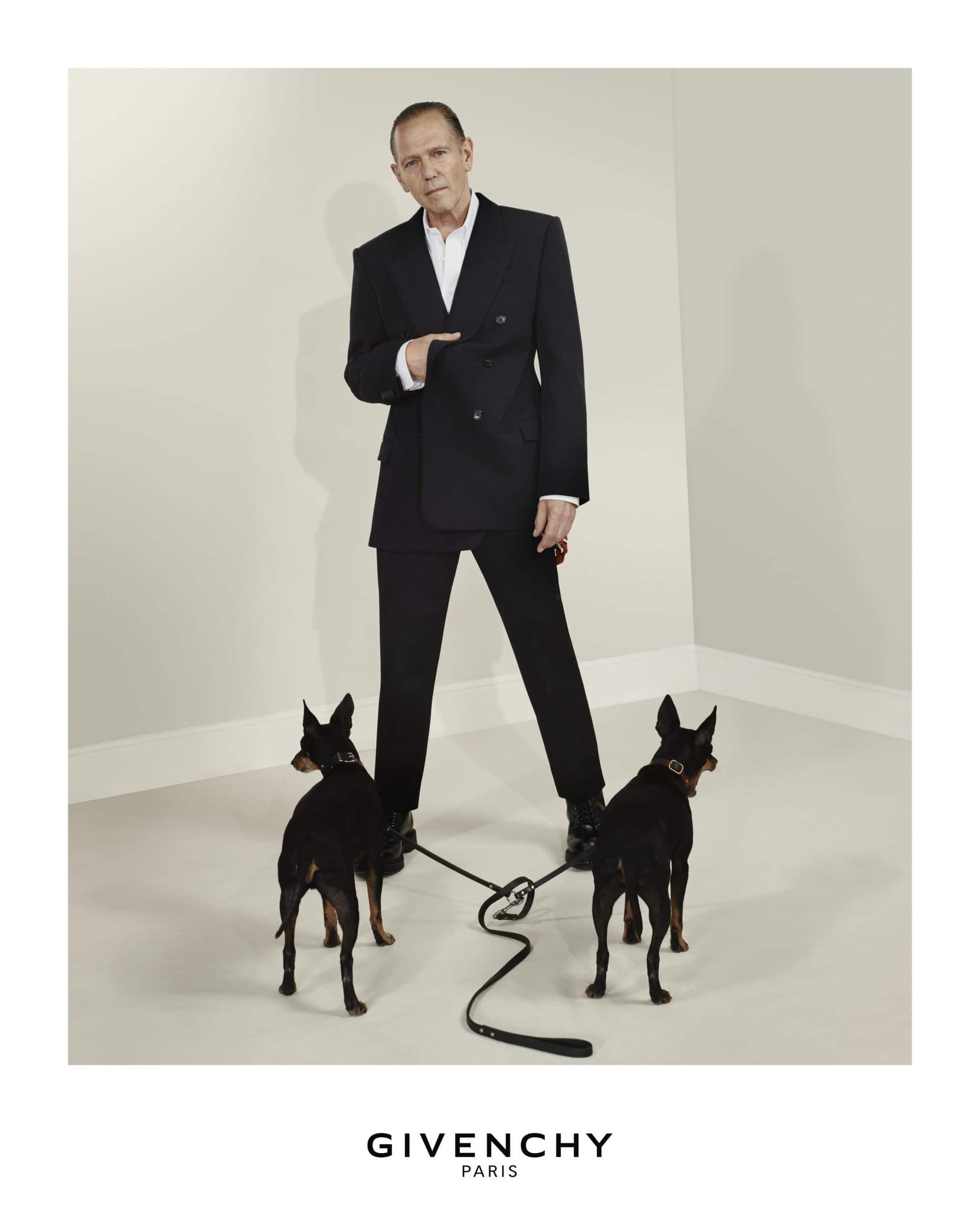 Stylish man in a black suit walking two black dogs on leashes in a minimalist studio, fashion forward and elegant with clean lines.