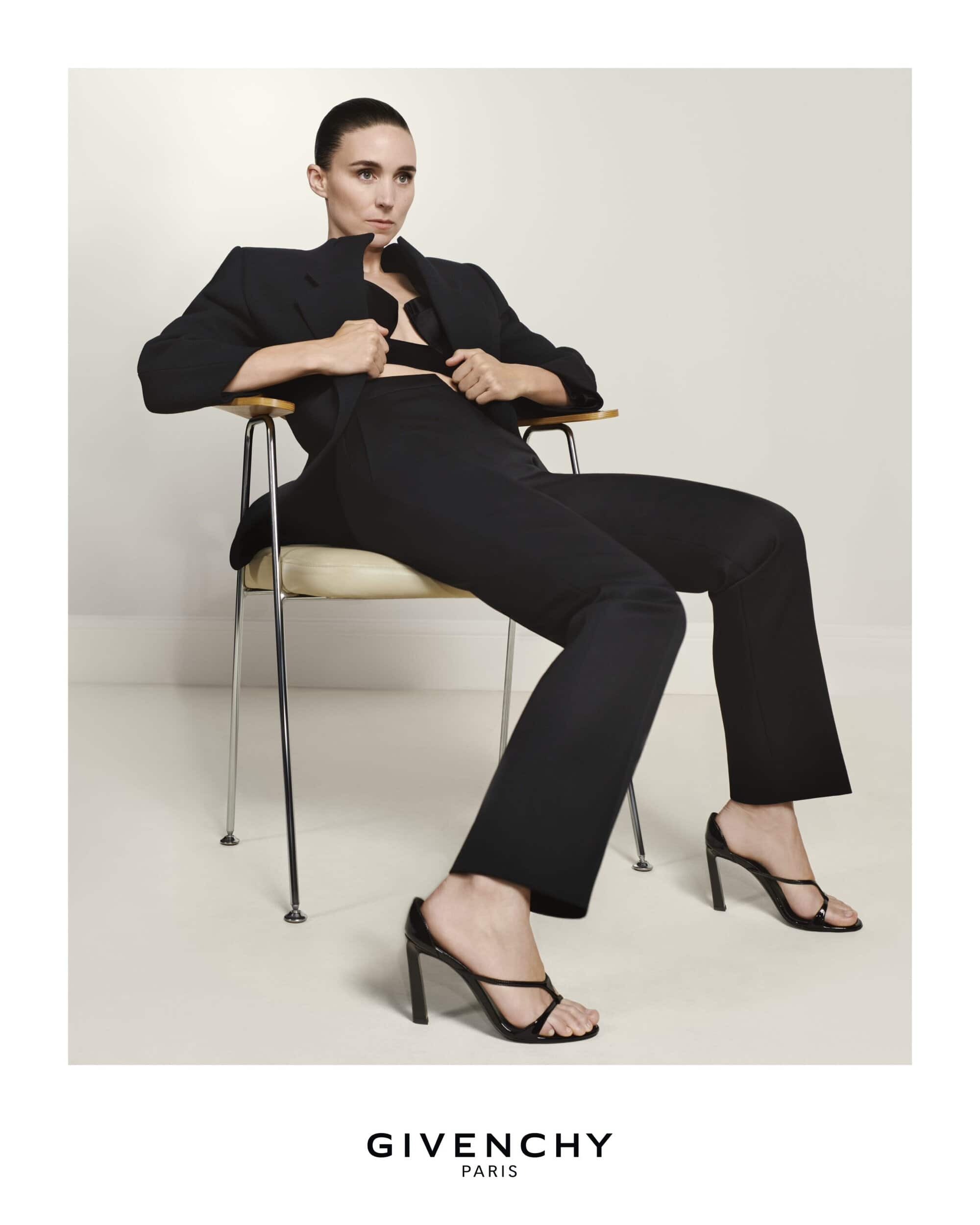 Rooney Mara seated in a modern chair wearing a black blazer and trousers against a pale background for Givenchy Spring 2026