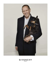 An older man holding a small black and white dog while wearing a dark jacket over a white shirt, set against a plain light background.