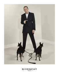 Man wearing a dark suit standing indoors with two black dogs on leashes on a light seamless background
