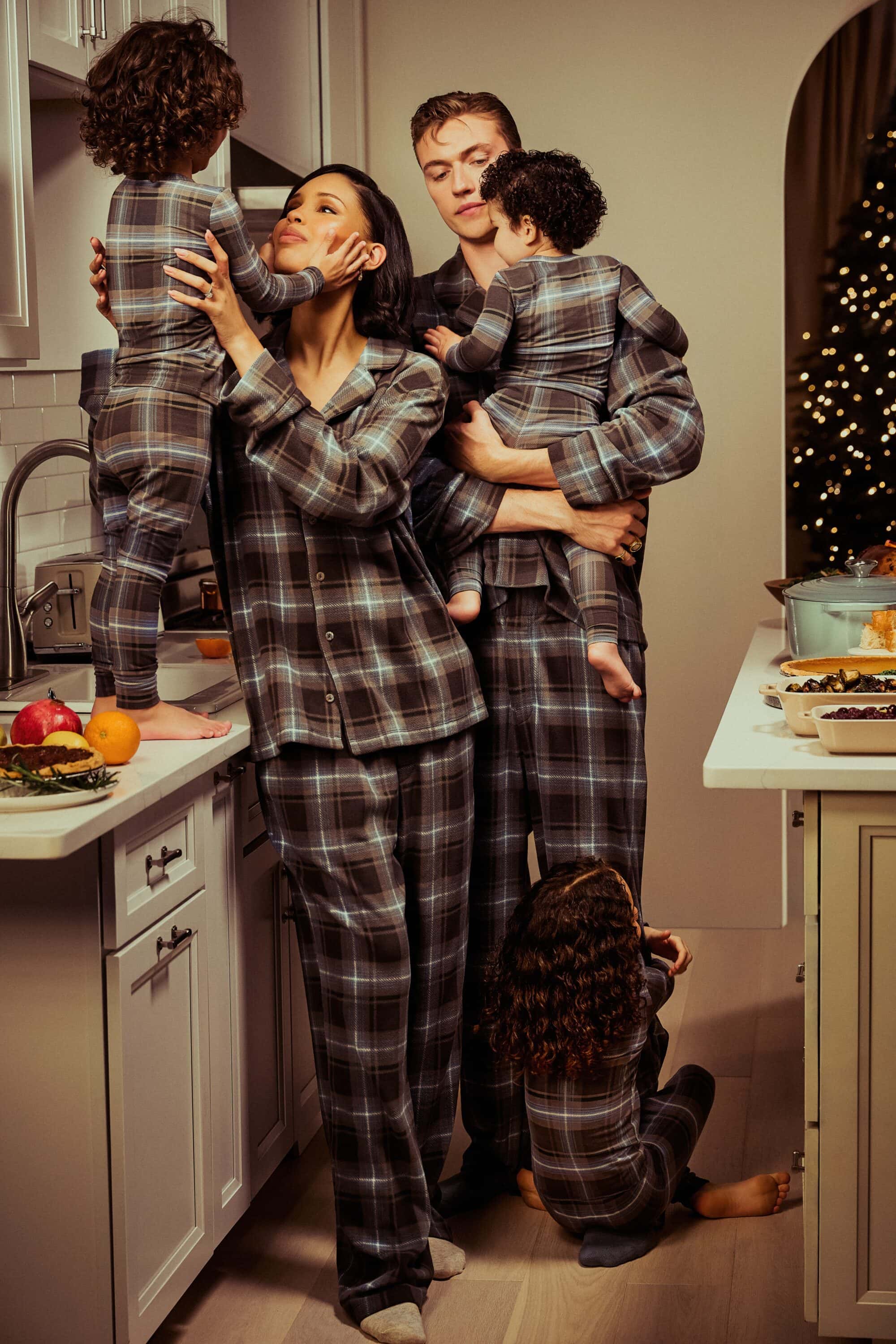 Family of four wears matching plaid pajamas in kitchen with Christmas tree. Mother holds child while other kids stand nearby.