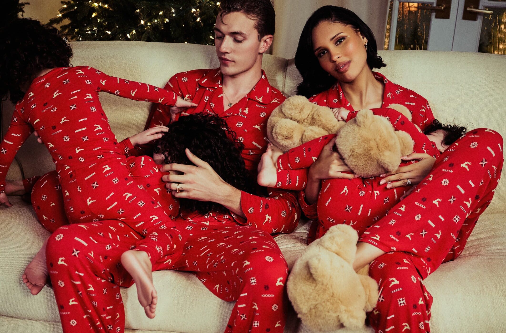 Family of five, including two adults and three children, in matching red Christmas pajamas sitting together on a light-beige couch indoors.