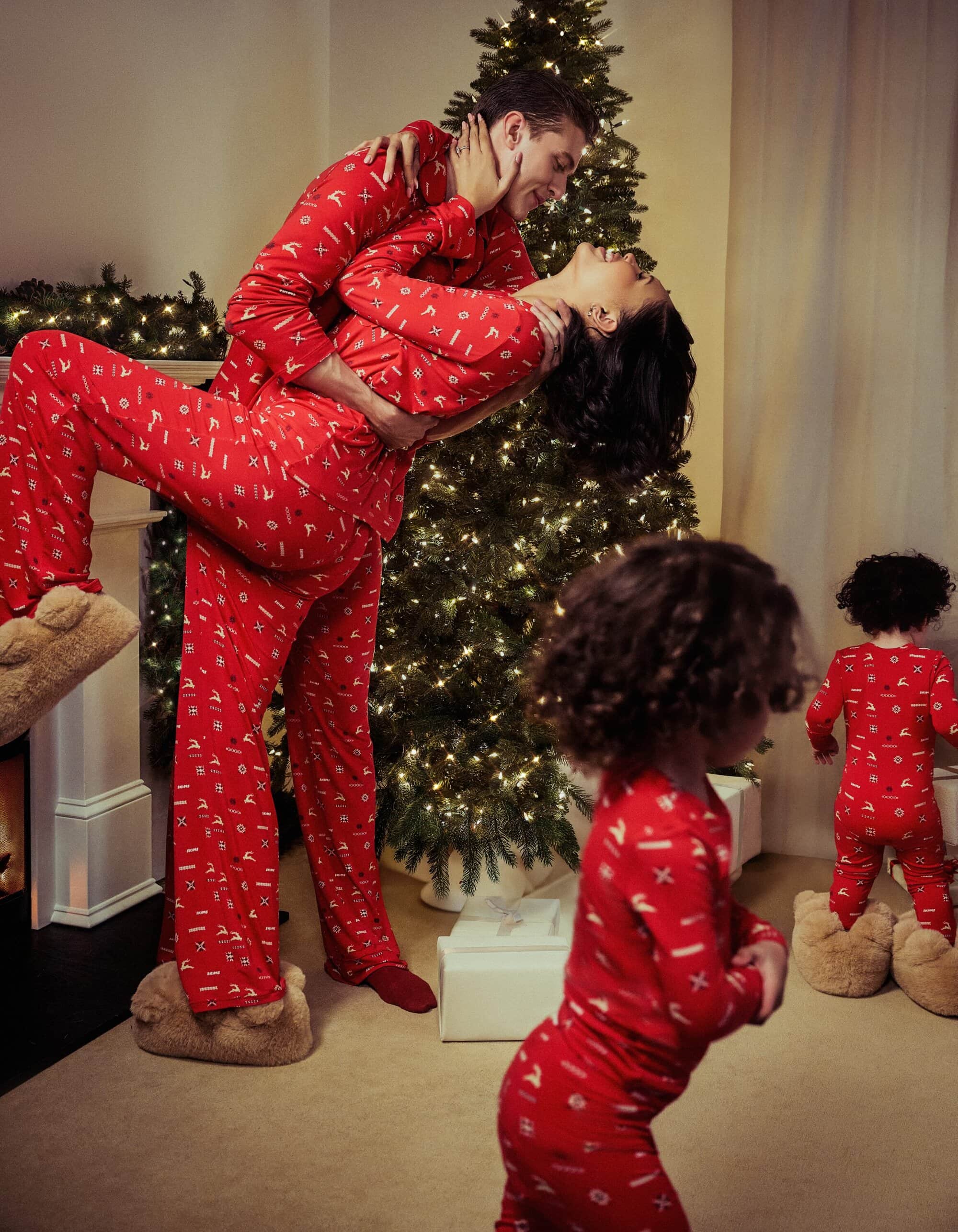A family of four, including a couple and two young children, wear matching red patterned pajamas indoors with a decorated Christmas tree.