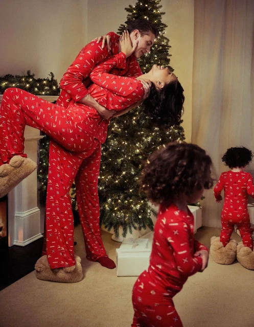 A family of four, including a couple and two young children, wear matching red patterned pajamas indoors with a decorated Christmas tree.