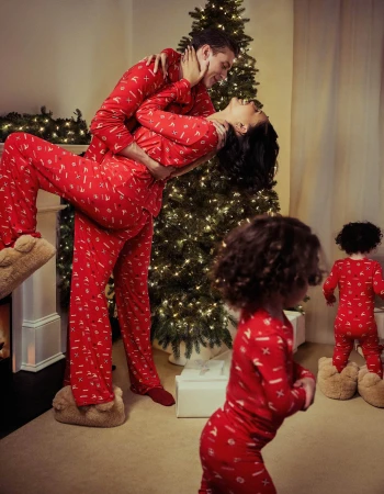 A family of four, including a couple and two young children, wear matching red patterned pajamas indoors with a decorated Christmas tree.