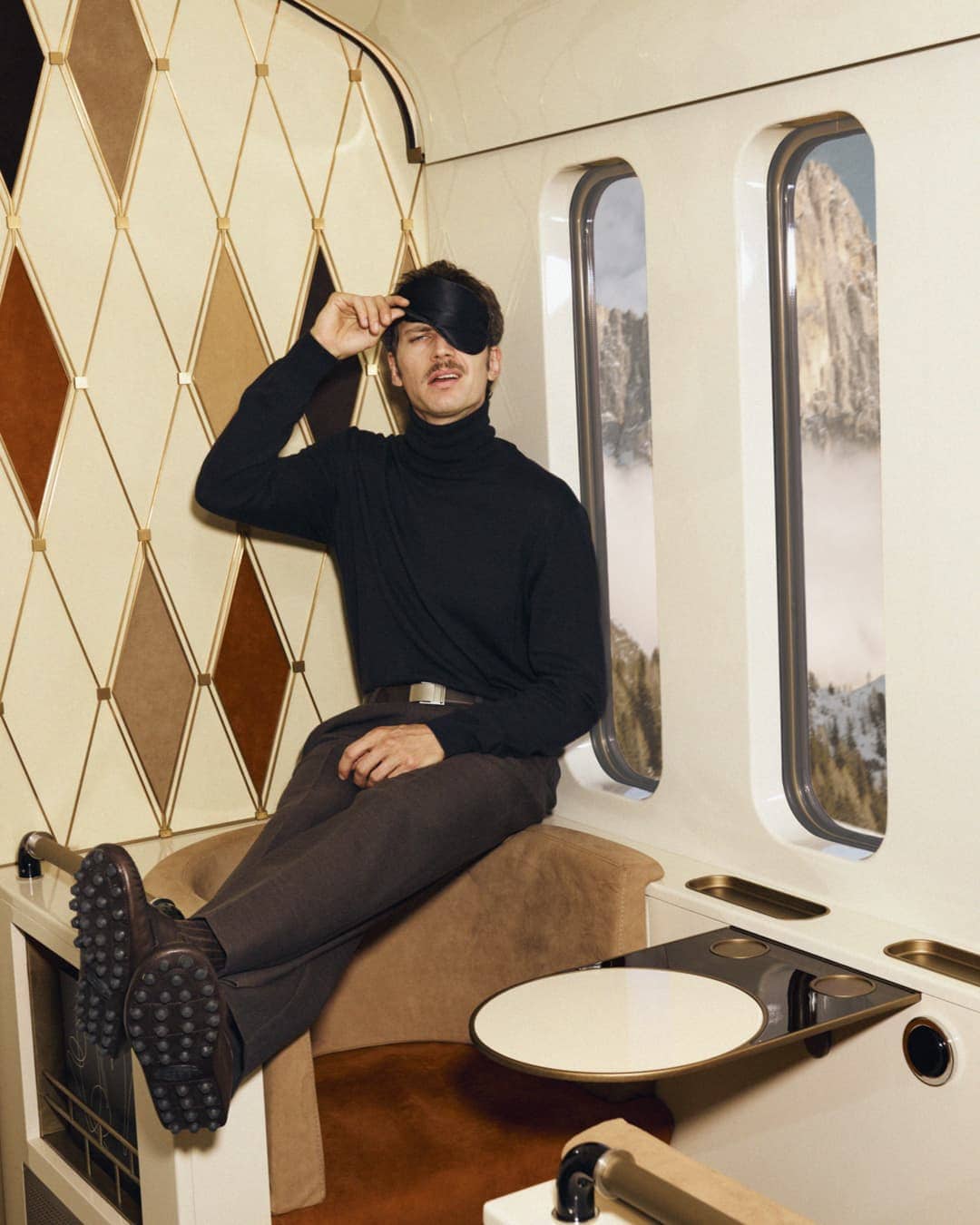 Adult male in turtleneck and brown trousers sits in a train car with mountain view, wearing sleep mask on his forehead.