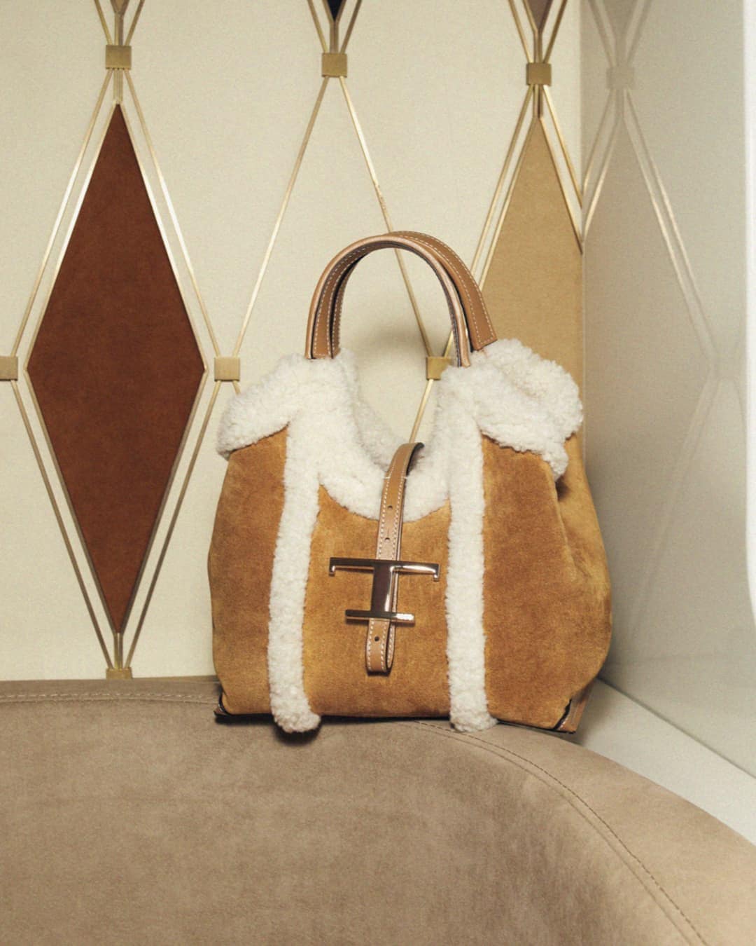 Tan suede Tod's handbag with cream shearling trim and leather handles on a beige suede chair against a geometric diamond wall.