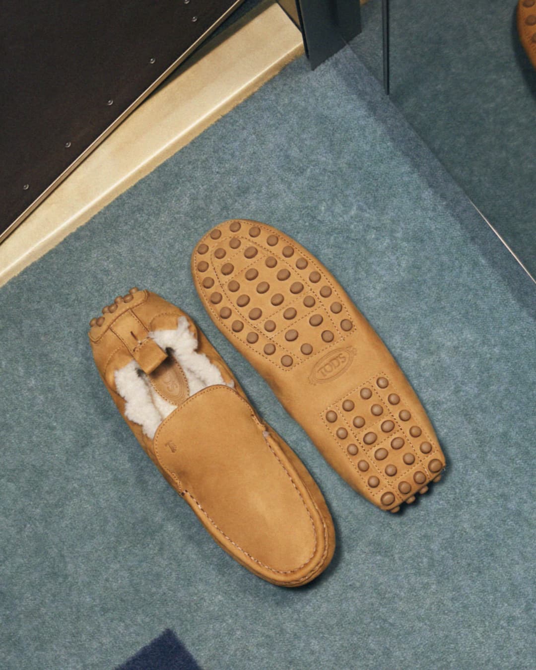 Tan leather Tod's loafers with white shearling interior rest on a blue carpet, one sole visible, in a product-focused shot.