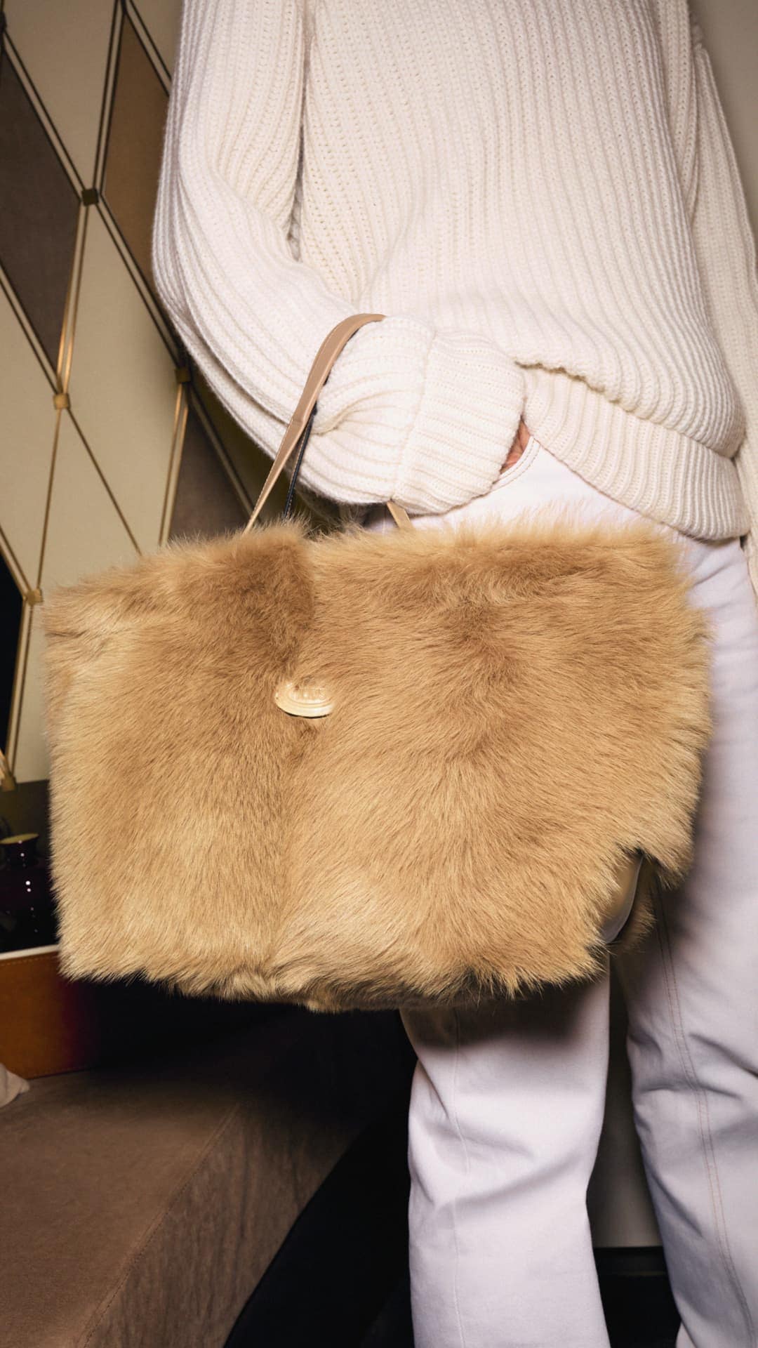A woman wearing a white sweater and pants holds a rectangular tan-colored furry bag with a light brown strap in an indoor setting.