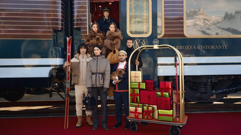 Group of people standing on a red carpet in front of a train with gifts, ski equipment, and dog, conveying a luxury travel theme.