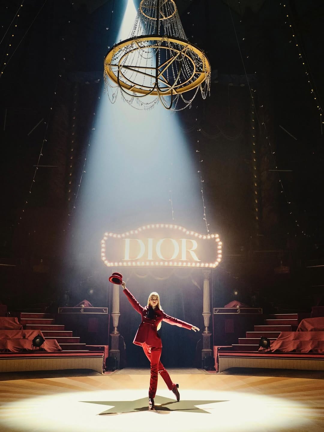 A woman in a red suit and hat is illuminated by a spotlight on a stage with a chandelier and signage for Dior.