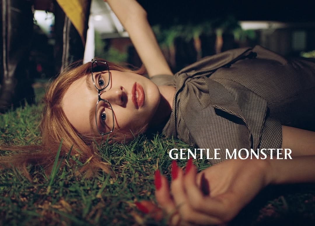 Adult female model with red hair, glasses, and red nails lying on green grass in an outdoor setting, wearing a patterned brown outfit.