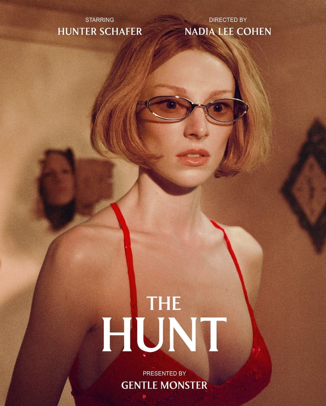 Portrait of Hunter Schafer wearing glasses and a red dress, against a textured wall with film credits in the poster for 'The Hunt'.