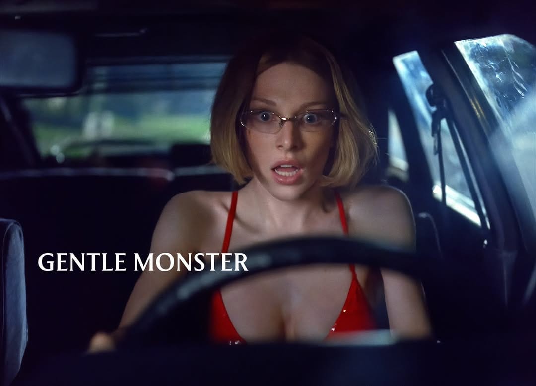 Woman in red top driving a car at night with a surprised expression and glasses on, with logo for Gentle Monster.