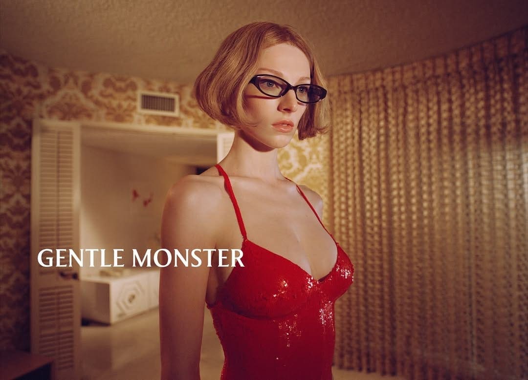 A woman with short blonde hair and glasses wears a red, sparkly dress in a staged photo studio environment with patterned wallpaper.