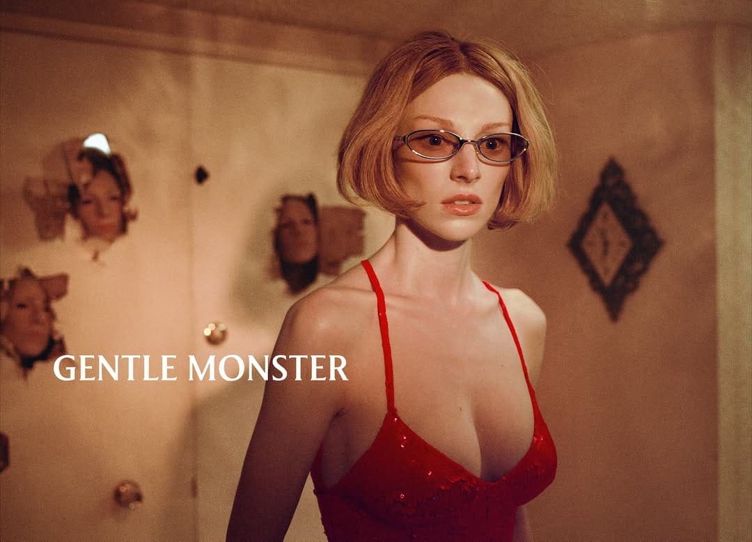 A woman with blonde hair and red dress wearing Gentle Monster glasses stands indoors with a wall of female faces behind her.