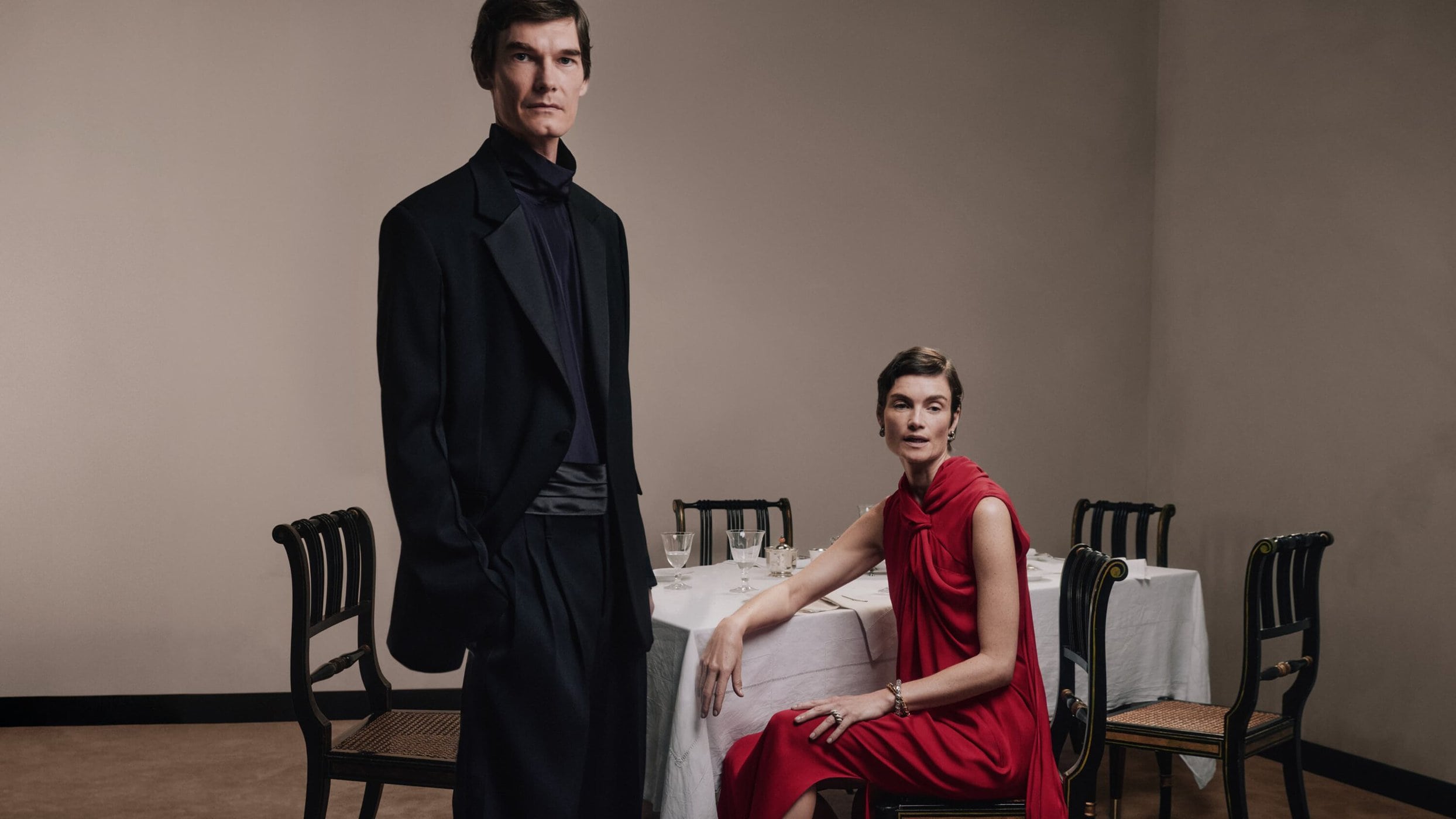 Man standing in dark suit and woman seated in red dress near a dining table in an interior setting for Lanvin.