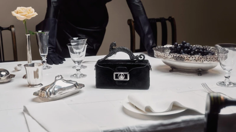 Close-up shot of black handbag with silver hardware on a white tablecloth set with silver tableware, glasses, grapes, and a rose.