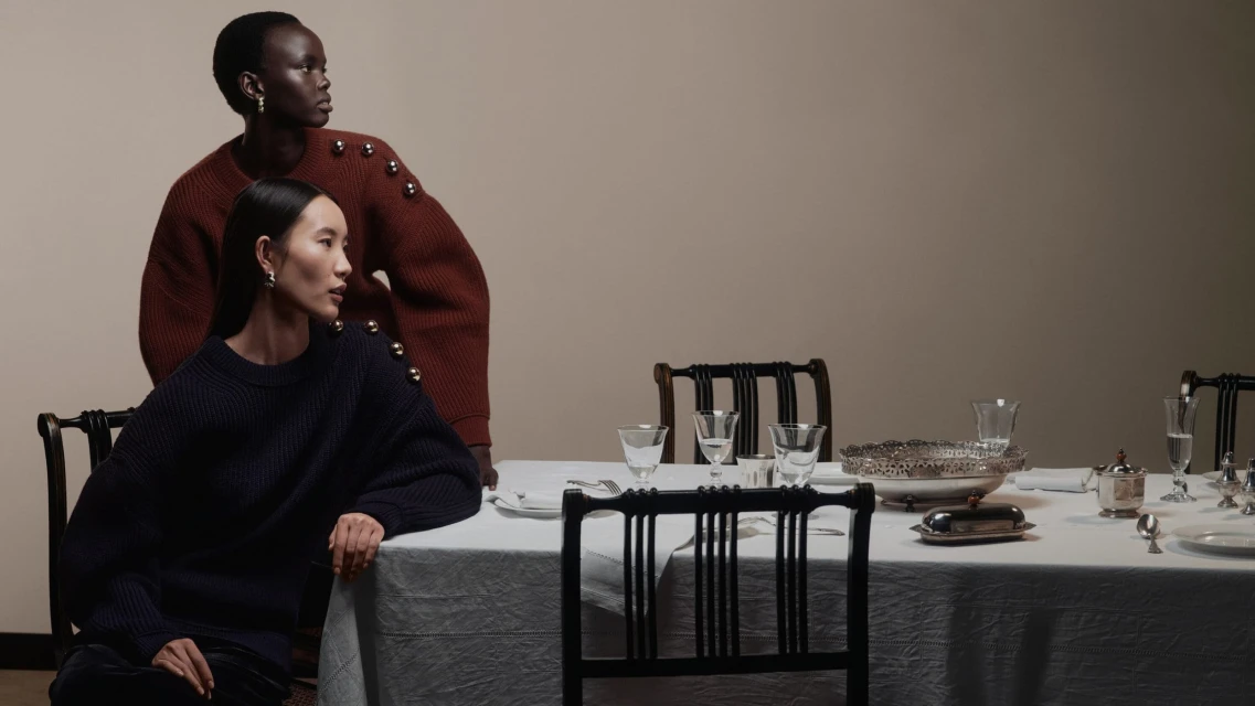 Two women in knit sweaters pose near a dining table with silver servingware and glassware in an indoor setting with neutral colors.