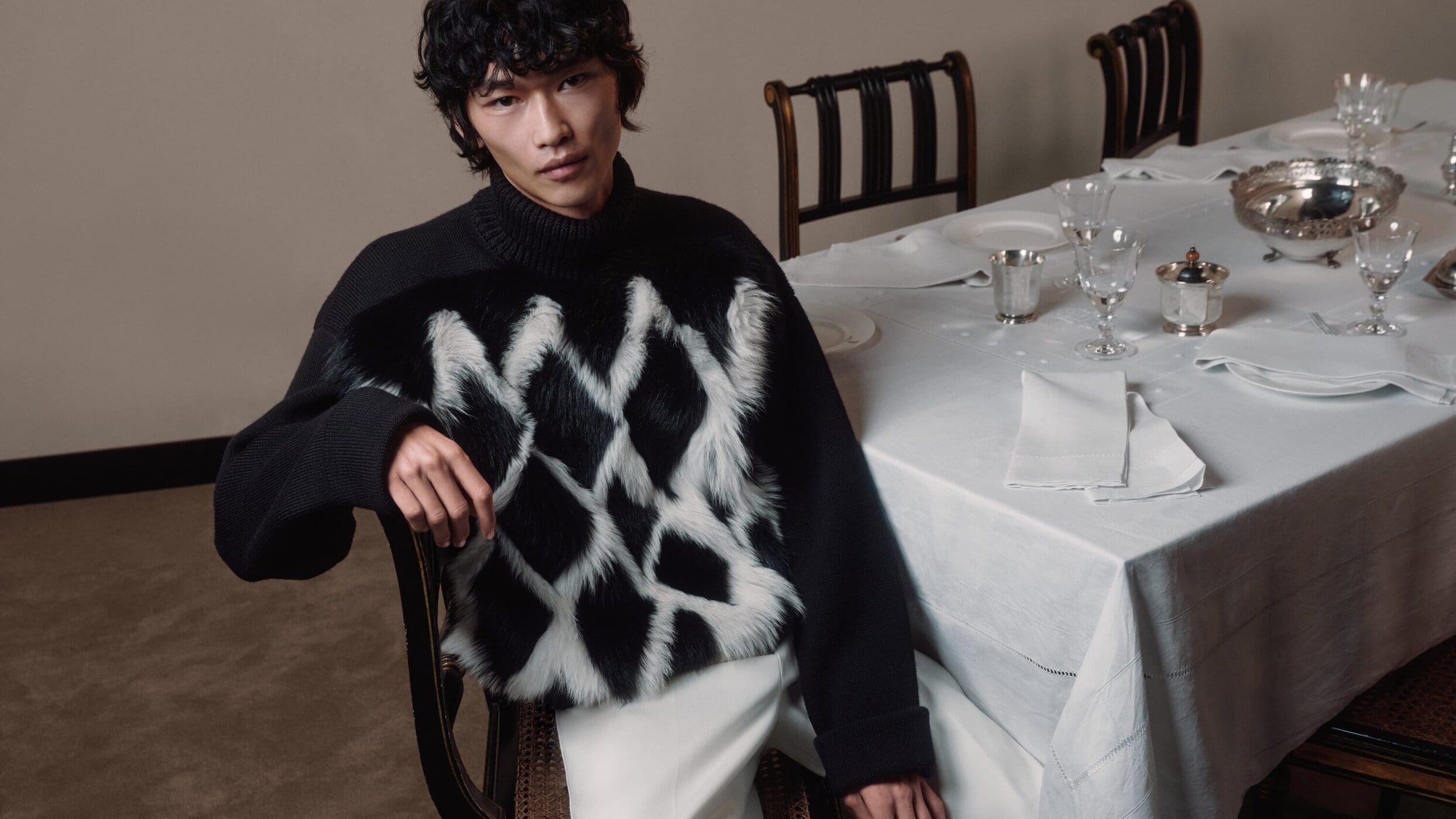 Asian man in a black sweater with white faux fur diamond pattern sitting at a table set with fine china and silver in a neutral interior.