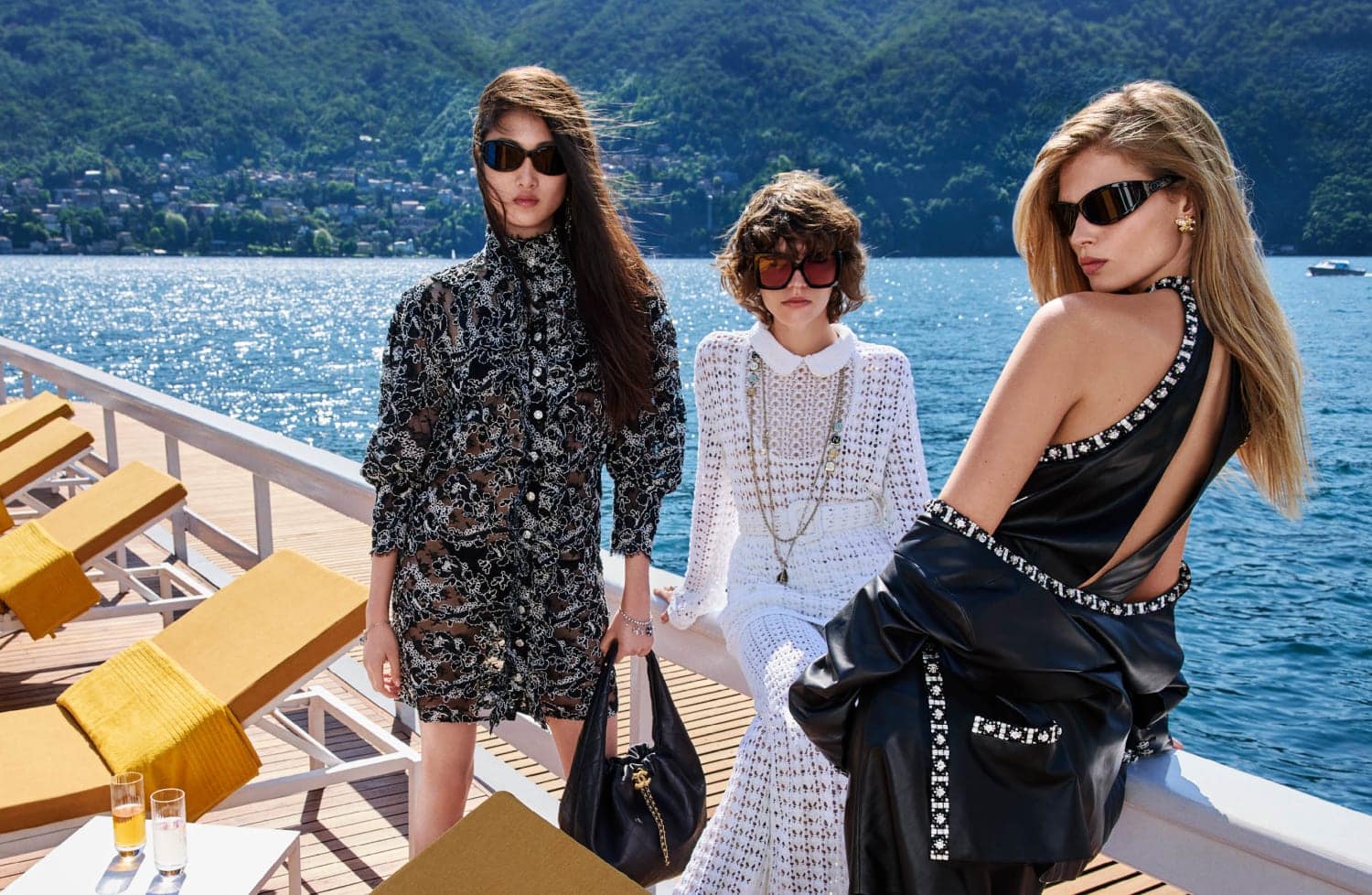 Three fashion models pose on a yacht deck wearing dresses and sunglasses with a background of a scenic lake and mountains.