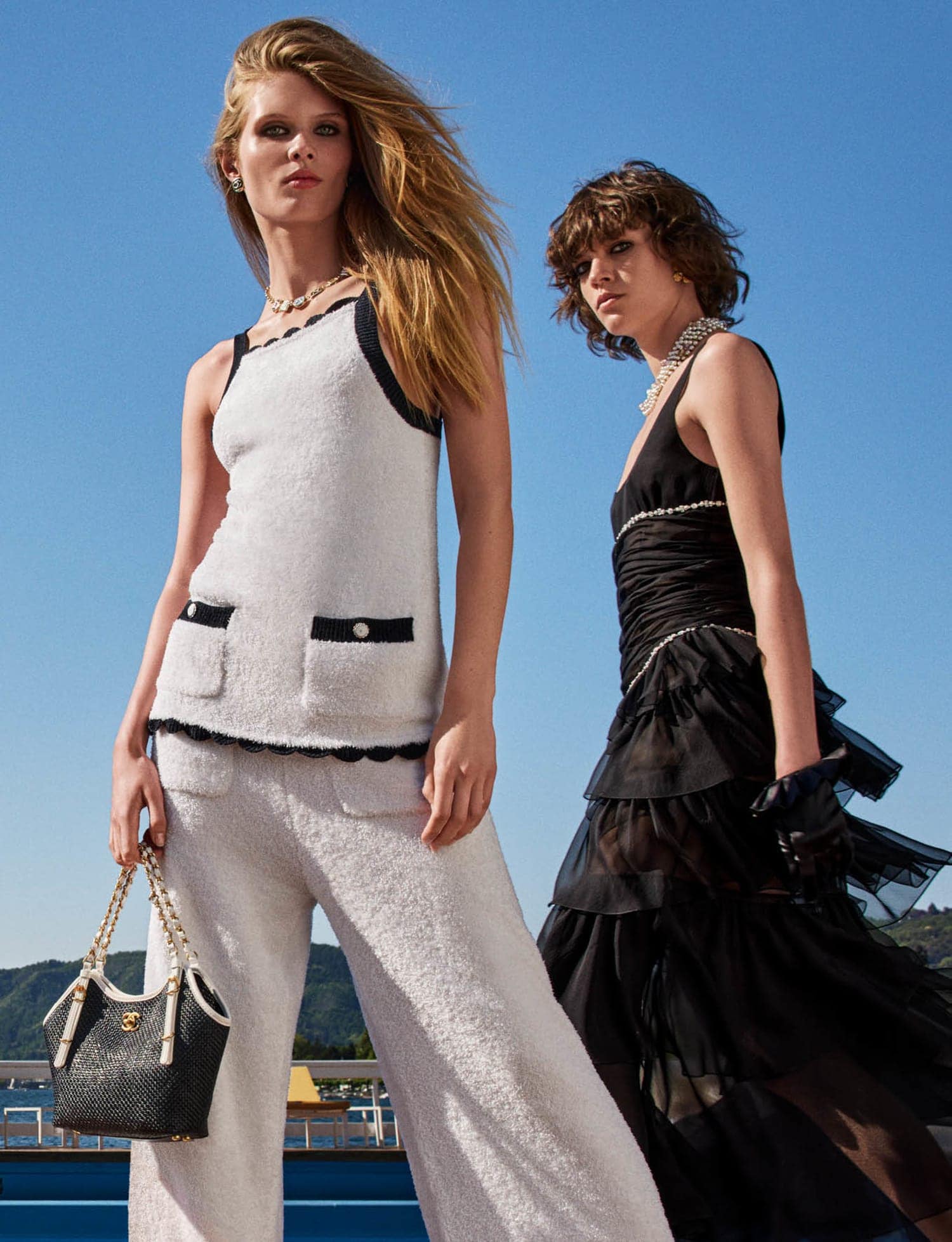 Two fashion models pose outdoors in white and black ensembles against a clear blue sky, showcasing elegant summer apparel and accessories.