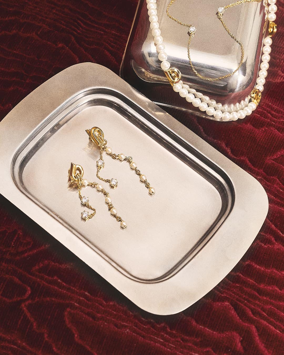 Elevated view of gold drop earrings with pearls on a silver tray, gold necklace with pearls, and ruby red velvet background.