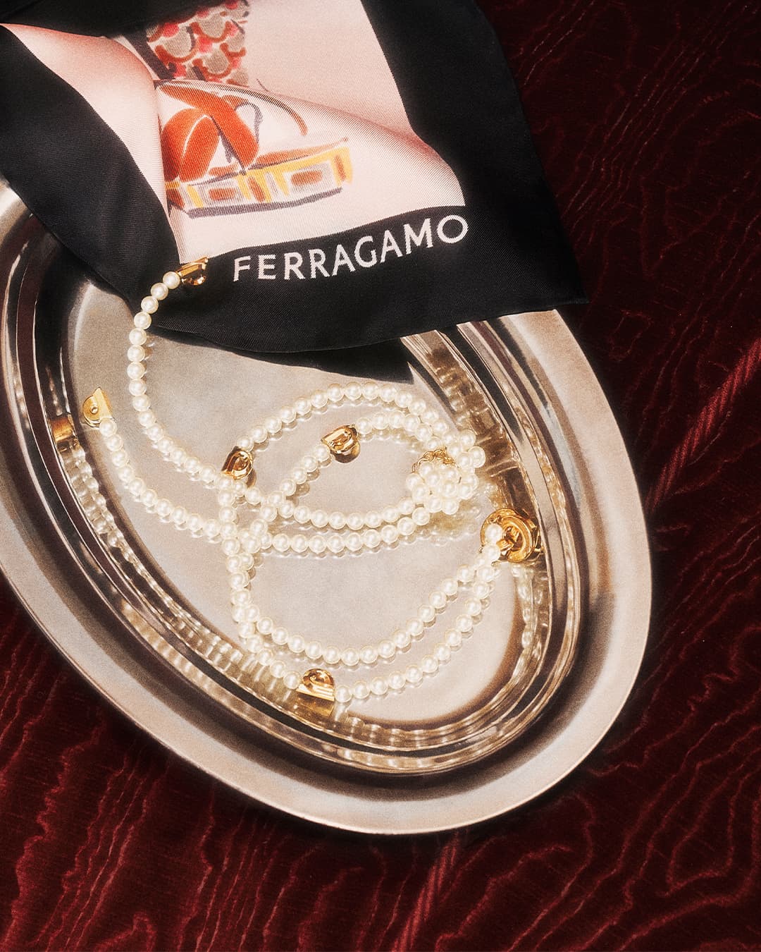Still life of a pearl necklace on a silver tray with a Ferragamo scarf featuring a shoe design and dark red velvet background.