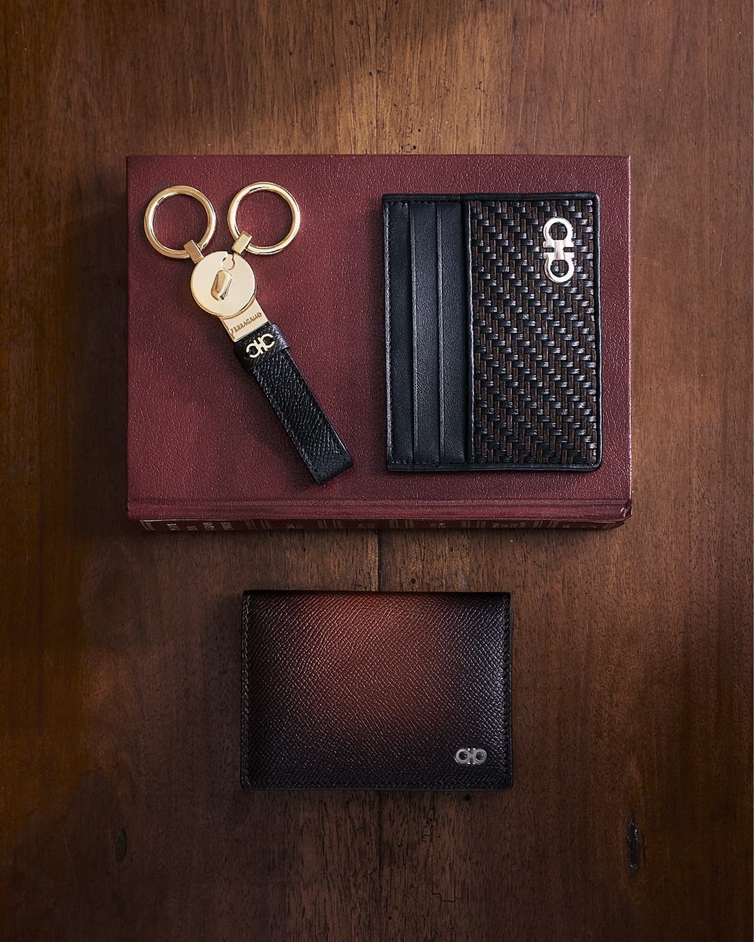 Elevated studio shot showcasing a brown wallet, a black card holder, and a black leather keyring with gold rings on a red book and wood surface.