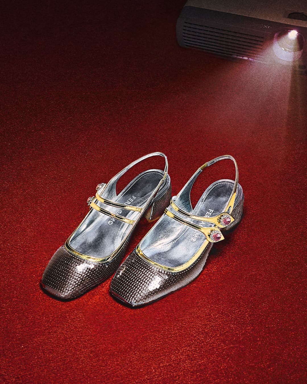 Sparkly Mary Jane heels with slingbacks sit on a red background with a projector light shining on the upper right corner.