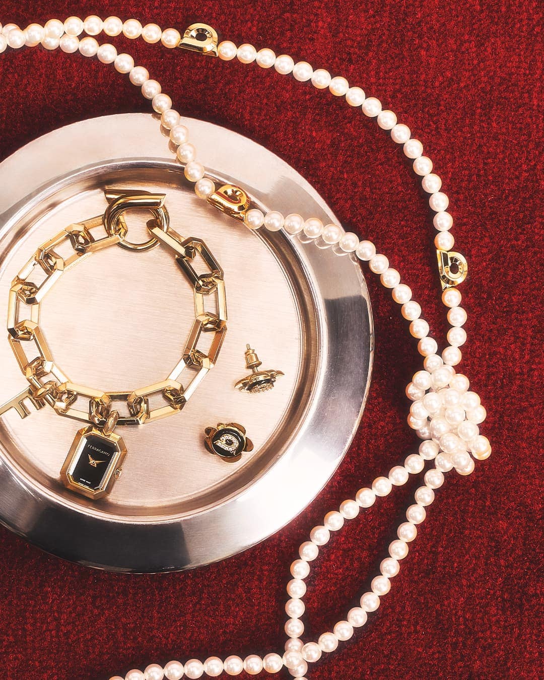 Still life of gold jewelry including a chain bracelet watch, earrings, and a pearl necklace displayed on a silver platter and red background.