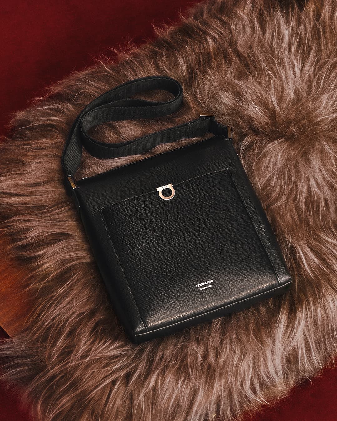 Elevated shot of a black leather messenger bag with a metal logo on a textured brown fur surface against a red background, product-focused.