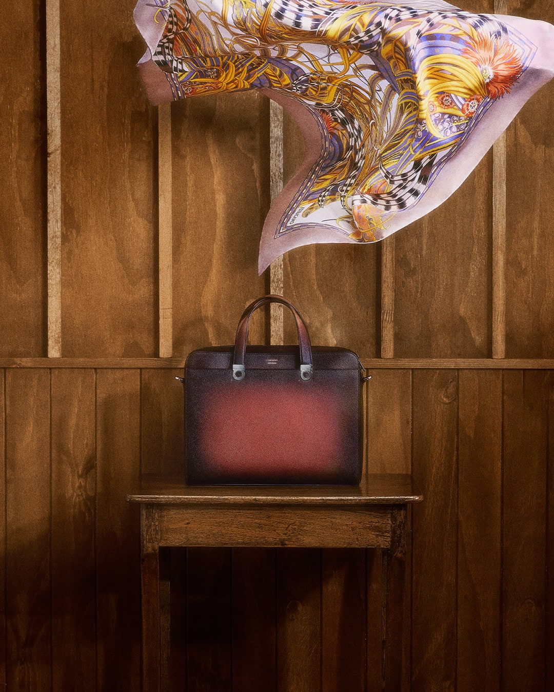 Stylish leather briefcase in brown and red tones on a wood table with a colorful scarf against a wooden wall. Fashionable product shot.