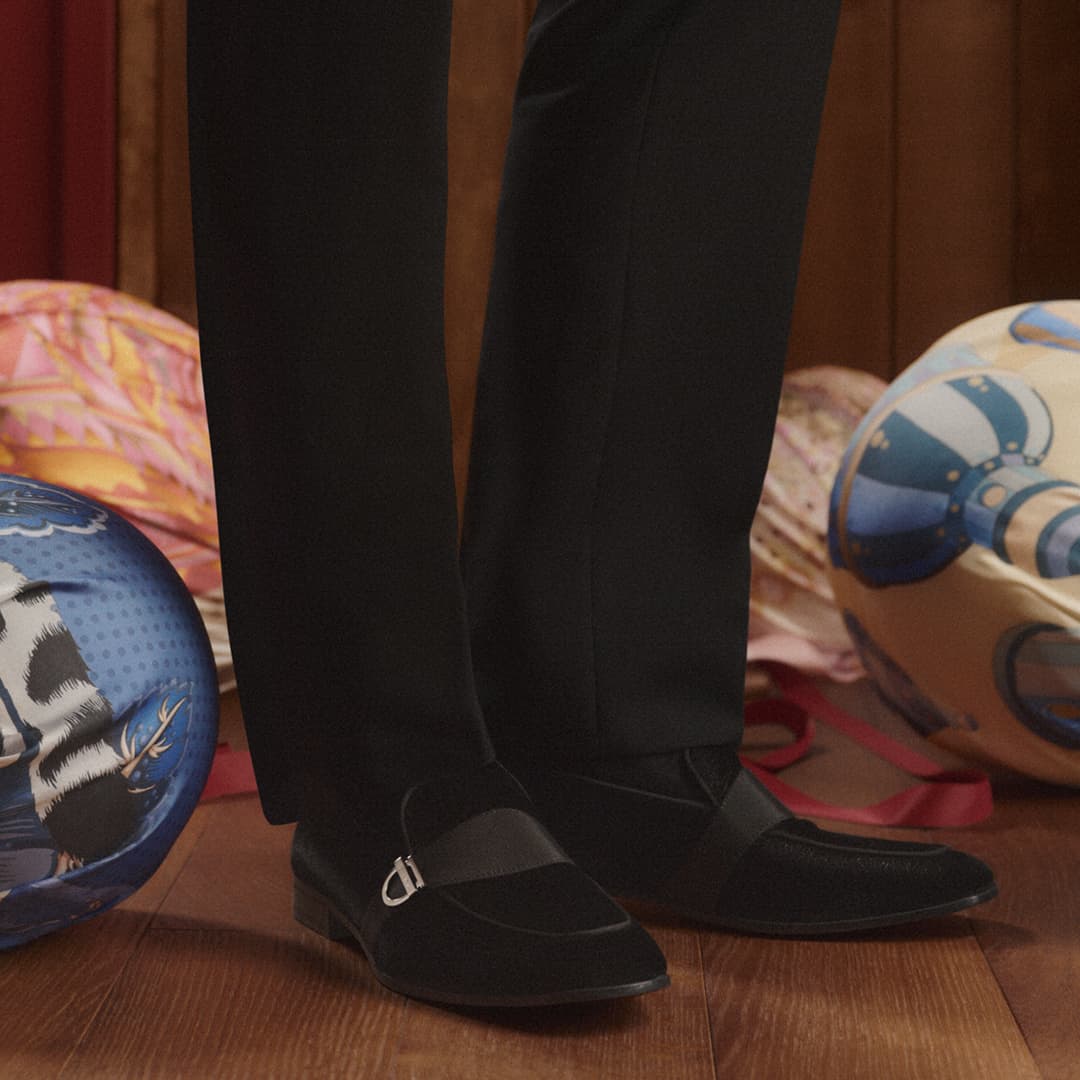 Black suede loafer shoes with metal detail worn with black dress pants stand on a wood floor, surrounded by patterned objects.