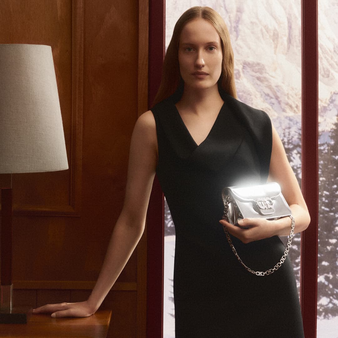 Indoor shot of a model wearing a black dress and holding a silver purse in front of a window with a snowy mountain landscape.