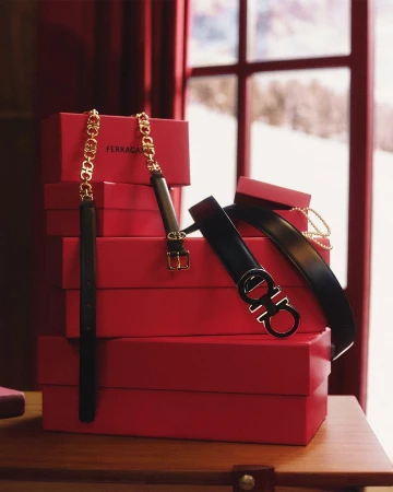 Ferragamo belts are displayed on top of a stack of red gift boxes, showcasing a minimalist aesthetic with soft lighting and an outdoor backdrop.